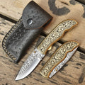 Open and closed pair of custom hand-engraved folding pocket knives with gold floral brass handles next to stamped dark leather sheath on wooden background