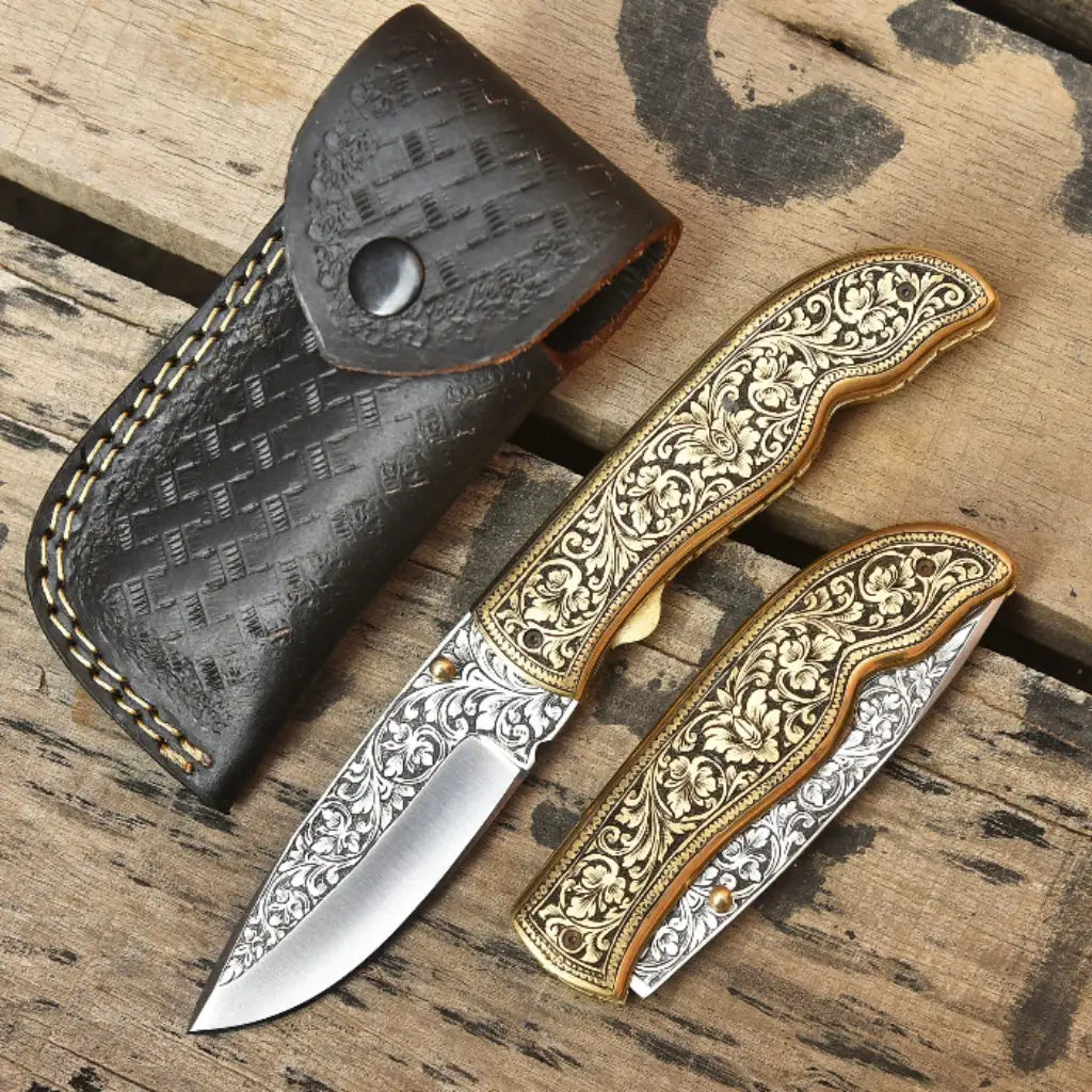 Open and closed pair of custom hand-engraved folding pocket knives with gold floral brass handles next to stamped dark leather sheath on wooden background