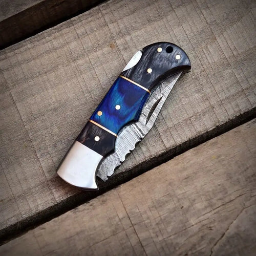 Closed handmade Damascus steel folding pocket knife with a dark blue wood handle resting on a wooden surface.