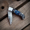Half-open handmade Damascus steel pocket knife showing filework blade detail and a blue resin wood handle.