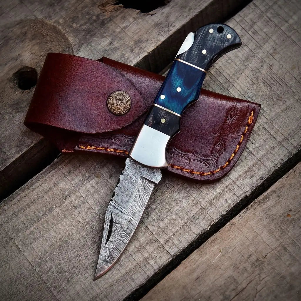 Handmade Damascus steel pocket knife with blue resin handle and custom brown leather sheath on wood background.